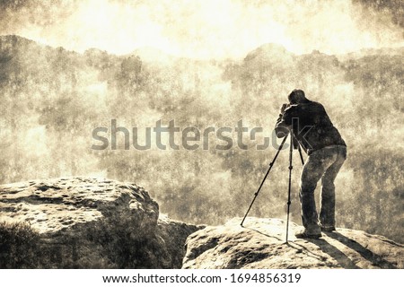 Professional photographer at tripod on cliff and hard working. Dreamy fogy landscape blue misty sunrise in a beautiful valley below Calotype filter. Old photo style.