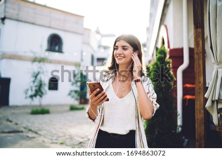 Happy Caucasian woman in electronic equipment dialing contact number for communicate on leisure, cheerful hipster girl in earphones listening positive music podcast from smartphone application