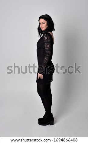 Portrait of a goth girl with dark hair wearing black dress and boots. Full length standing pose in life profile on a studio background.
