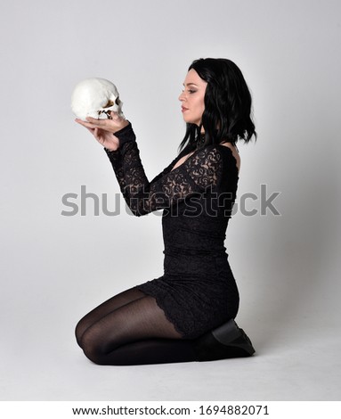 Portrait of a goth girl with dark hair wearing black dress and boots. Full length sitting pose on a studio background
