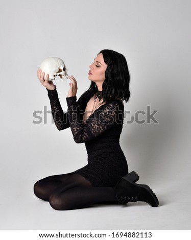 Portrait of a goth girl with dark hair wearing black dress and boots. Full length sitting pose on a studio background