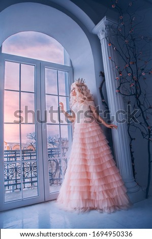 Fantasy beautiful medieval woman princess stands near window castle white room. Girl looking outdoor with hope . Pink long luxury dress. Lady queen wavy blonde hair. Vintage royal crown. Stay home 