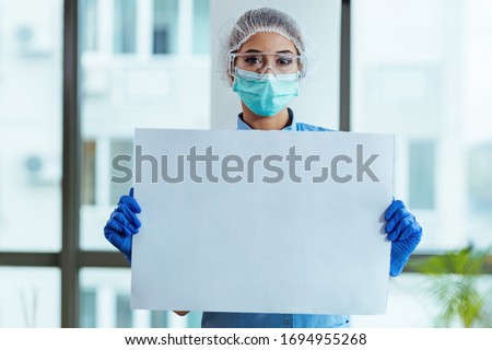 Female doctor with protective workwear holding empty cardboard while standing at clinic and looking at camera. Copy space.
