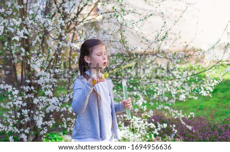 Beautiful little girl, has happy face, pretty eyes, brown hair, dressed in white t-shirt and gray jacket, playing soap bubbles in flowering trees. Child portrait. Spring view. Kids fashion style. 