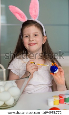 Beautiful little brunette girl, has happy fun smiling face, pretty eyes, white t-shirt, hare ears, paint Easter eggs. Child portrait and kids hobby concept. Holiday accessories. 