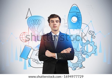 Portrait of confident young European businessman standing with crossed arms near gray wall with creative blue startup sketch drawn on it.