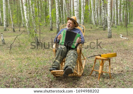 Caucasian senior wearing straw hat singing and playing garmonika while sitting lonely in wicker rocking chair in birch forest