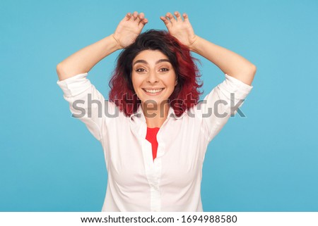 I'm rabbit! Portrait of playful childish woman with fancy red hair in white shirt gesturing bunny ears on her head and smiling carefree, having fun. indoor studio shot isolated on blue background