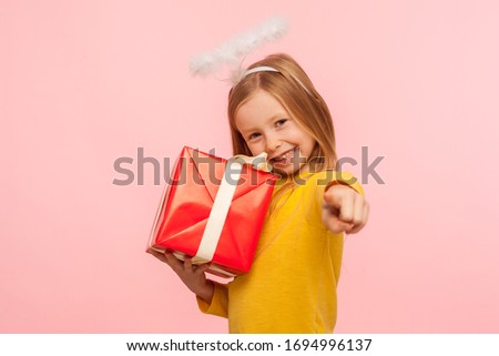 Hey, this present for you! Portrait of happy little ginger girl with angelic halo embracing wrapped box and pointing to camera, choosing lucky winner to give bonus, gift. indoor studio shot, isolated