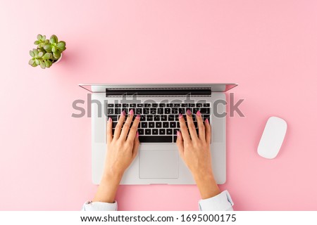Female hands using laptop on pink background with computer mouse and flower. Office desktop. Top view