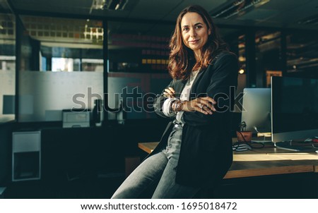 Mature businesswoman sitting on her desk. Confident female entrepreneur with her arms crossed looking at camera.