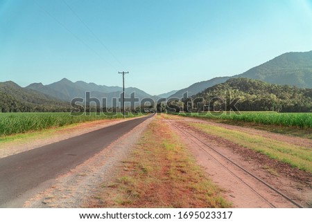 Road journey & Beautiful mountain landscape with sugar cane fields foreground. Dramatic view of rail, fields, trees, green forest, farm, mountains, blue sky & road. Walsh's Pyramid, Cairns, Australia.