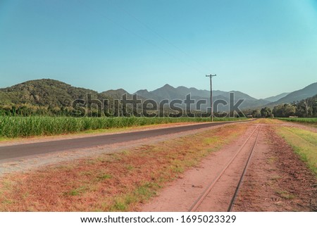 Road journey & Beautiful mountain landscape with sugar cane fields foreground. Dramatic view of rail, fields, trees, green forest, farm, mountains, blue sky & road. Walsh's Pyramid, Cairns, Australia.