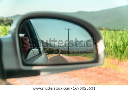 Rearview car mirror reflection of Beautiful mountain landscape with sugar cane fields foreground. Road, fields, trees, green forest, farm, mountains, blue sky & road. Walsh's Pyramid, Cairns Australia