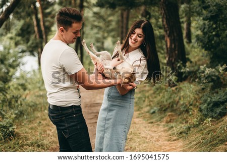 Young white couple holding a small corgi dog in the summer forest in nature. Woman in white shirt, man in white t-shirt and jeans. Happy family, fun, europe, romance, hugs, tenderness.