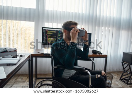 Male programmer in a wheelchair wearing virtual reality glasses while working at his workplace with multiple computer screens on the background. Disability concept. IT software. High Tech