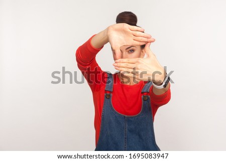 Capture moments. Portrait of girl in denim overalls looking through photo frame shape with fingers, viewing distant, focusing and zooming at camera. indoor studio shot isolated on white background