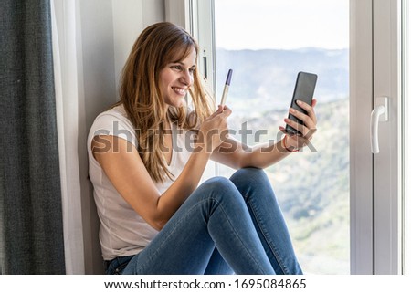 Young woman making a video call showing her pregnancy test to her partner with her mobile phone at home. Concept of new technologies and social networks at home.