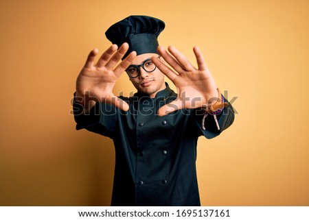 Young brazilian chef man wearing cooker uniform and hat over isolated yellow background doing frame using hands palms and fingers, camera perspective