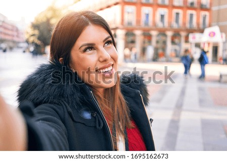 Young beautiful brunette woman smiling happy and confident. Standing with smile on face making selfie by the camera at street of city