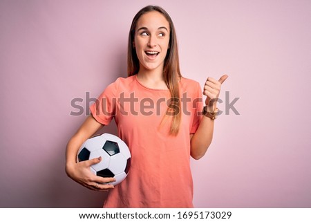 Young beautiful player woman playing soccer holding football ball  over pink background pointing and showing with thumb up to the side with happy face smiling