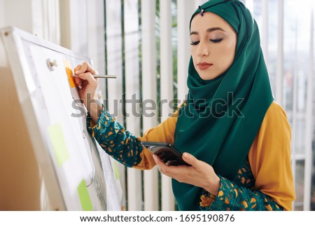 Young pretty muslim businesswoman in traditional clothes checking smartphone when writing ideas on colorful stickers