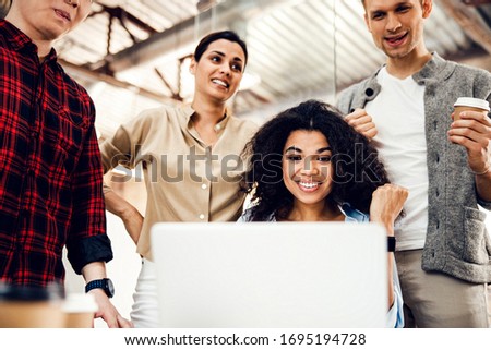Smiling young woman sitting at the table with notebook while coworkers standing beside her stock photo