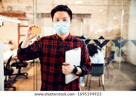 Good-looking Asian guy in protective antiviral mask holding bank card and laptop stock photo