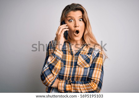 Beautiful blonde woman having conversation talking on the smartphone over white background scared in shock with a surprise face, afraid and excited with fear expression