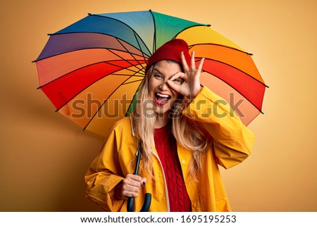 Young beautiful blonde woman wearing raincoat and wool cap holding colorful umbrella with happy face smiling doing ok sign with hand on eye looking through fingers