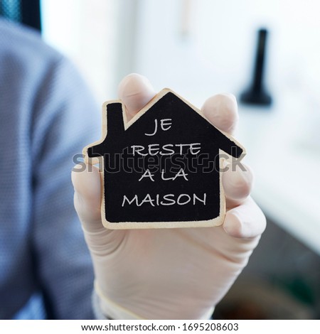 closeup of a man, in casual wear wearing latex gloves indoors, showing the message I stay at home in french written in a house-shaped black sign, as a measure to stop the spreading of the covid-19