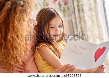 Traditional womans day concept. Young mother holding greeting cards from her little child girl, hugging daughter and sitting at home