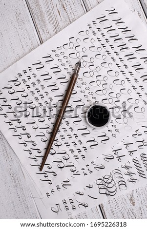 Calligraphy sheets, nibs, paper, ink on a wooden background. Letters of the English alphabet written with a paint brush.