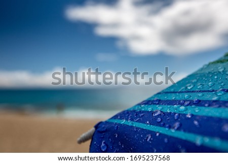 Close up rain drops on a blue striped sunbed part, blurred coastline clear blue cloudy sky on background, macro photography
