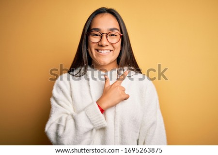 Young beautiful asian woman wearing casual sweater and glasses over yellow background cheerful with a smile of face pointing with hand and finger up to the side with happy and natural expression