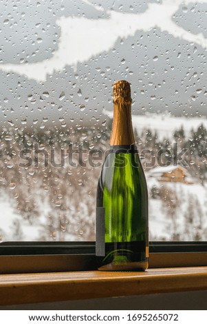 Champagne bottle on windowsill through the glass in the raindrops over mounains winter background.
