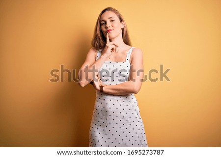 Young beautiful blonde woman on vacation wearing summer dress over yellow background looking confident at the camera smiling with crossed arms and hand raised on chin. Thinking positive.