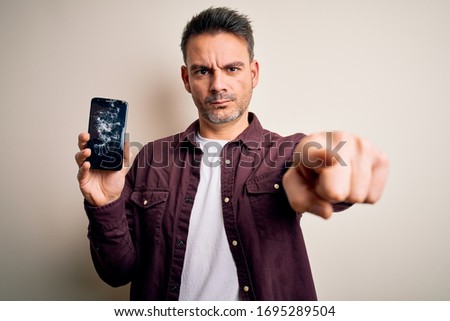 Young handsome man holding broken smartphone showing craked screen pointing with finger to the camera and to you, hand sign, positive and confident gesture from the front