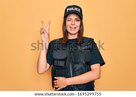 Young beautiful brunette policewoman wearing police uniform bulletproof and cap smiling with happy face winking at the camera doing victory sign with fingers. Number two.