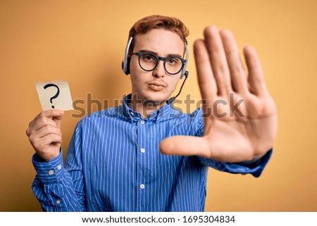 Redhead call center agent man working using headset holding reminder with question mark with open hand doing stop sign with serious and confident expression, defense gesture