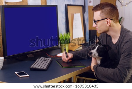 The dog sits in the man’s hands. A man works at home, a graphic designer, a creator works at home on freelance with a dog. The pet makes it difficult to work at home. A man is looking at green screen.