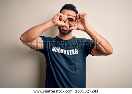 Handsome man with beard wearing t-shirt with volunteer message over white background Doing heart shape with hand and fingers smiling looking through sign