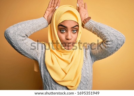 Young beautiful african american girl wearing muslim hijab over isolated yellow background Doing bunny ears gesture with hands palms looking cynical and skeptical. Easter rabbit concept.