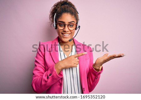 Young african american call center agent girl wearing glasses working using headset amazed and smiling to the camera while presenting with hand and pointing with finger.