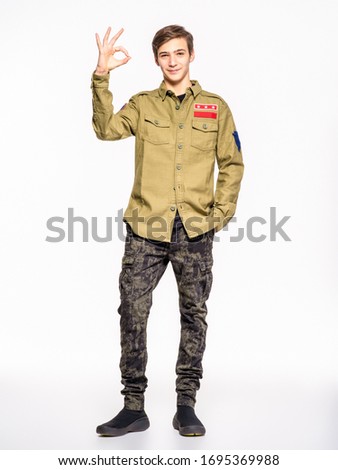 Young man in camouflage clothing shows big thumbs up. Rookie in army clothes. Teenager in khaki military clothing poses in a studio on a white background. Conscript in camouflage is showing big thumb.