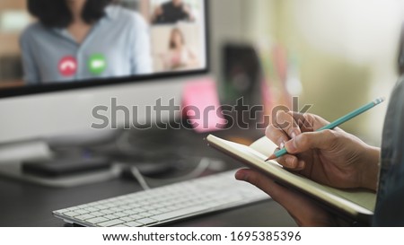 Closeup man taking note and sitting in front his computer monitor while video calling with colleague. Working from home through video conference concept. Royalty-Free Stock Photo #1695385396
