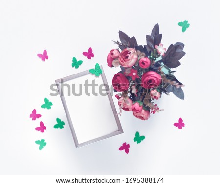 A metal photo frame and a bouquet of flowers are on a white background. Nearby are figures of butterflies made of felt. Minimalist arrangement. Flat lay. Copy space.