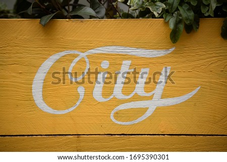 Vintage hand-lettered yellow painted sign on wood of the word, "City" with green plants peeking over the top.