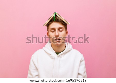 Portrait of a sleepy young man on a pink background with his eyes closed and a book on his head isolated on a pink background. Tired student boy stands on pink background. Copy space