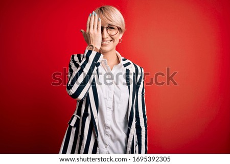 Blonde business woman with short hair wearing glasses and striped jacket over red background covering one eye with hand, confident smile on face and surprise emotion.